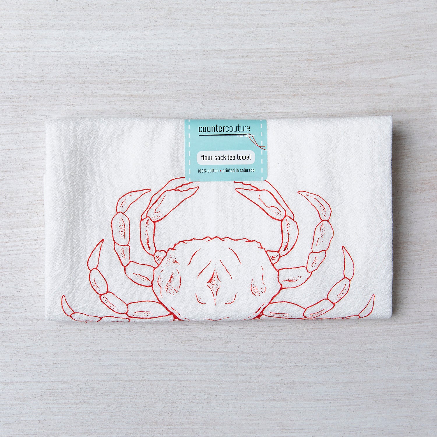 Folded Dungeness Crab flour sack tea towel by Counter Couture, displaying a red crab design with a tag indicating 100% cotton, perfect for kitchen use.