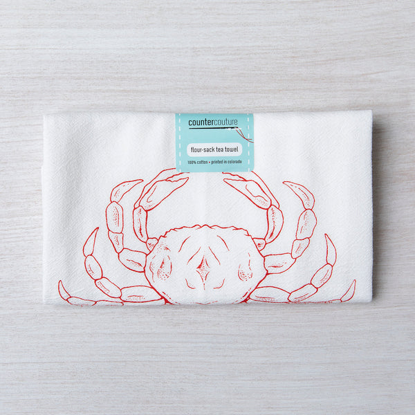 Folded Dungeness Crab flour sack tea towel by Counter Couture, displaying a red crab design with a tag indicating 100% cotton, perfect for kitchen use.