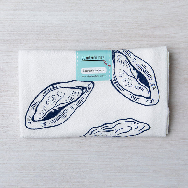 Folded oyster print flour sack tea towel by Counter Couture, showcasing detailed navy blue illustrations of oysters with a tag indicating material and design details.