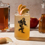 Sasquatch can glass filled with iced apple cocktail, apple slices garnish, apples and decanters nearby