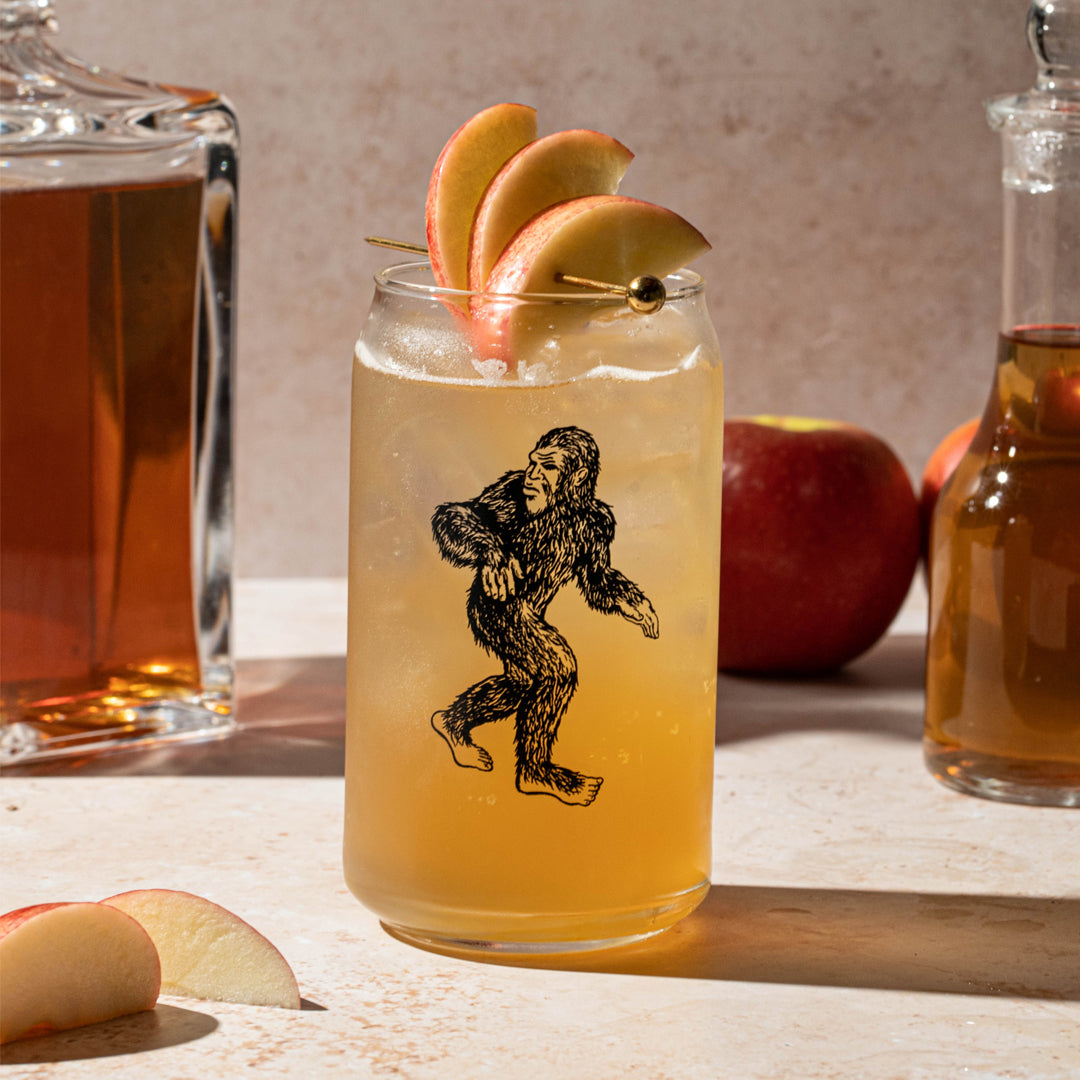 Sasquatch can glass filled with iced apple cocktail, apple slices garnish, apples and decanters nearby