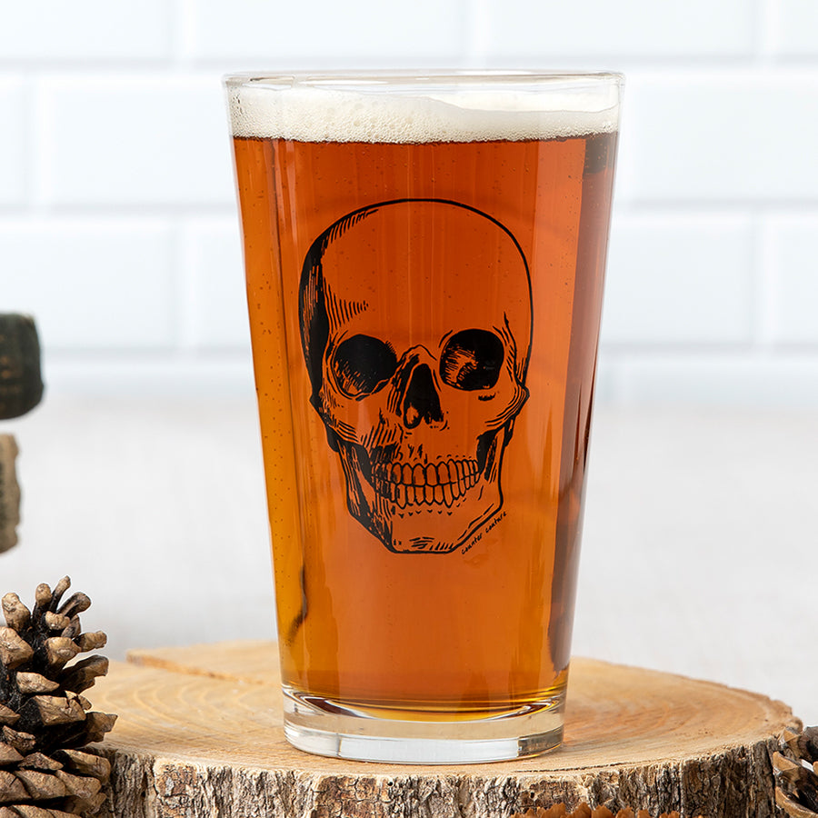 Pint glass of amber beer with black skull illustration, on wood coaster, rustic barware