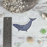 Blue whale vinyl sticker on textured background with seashells, sea glass, and ruler for scale