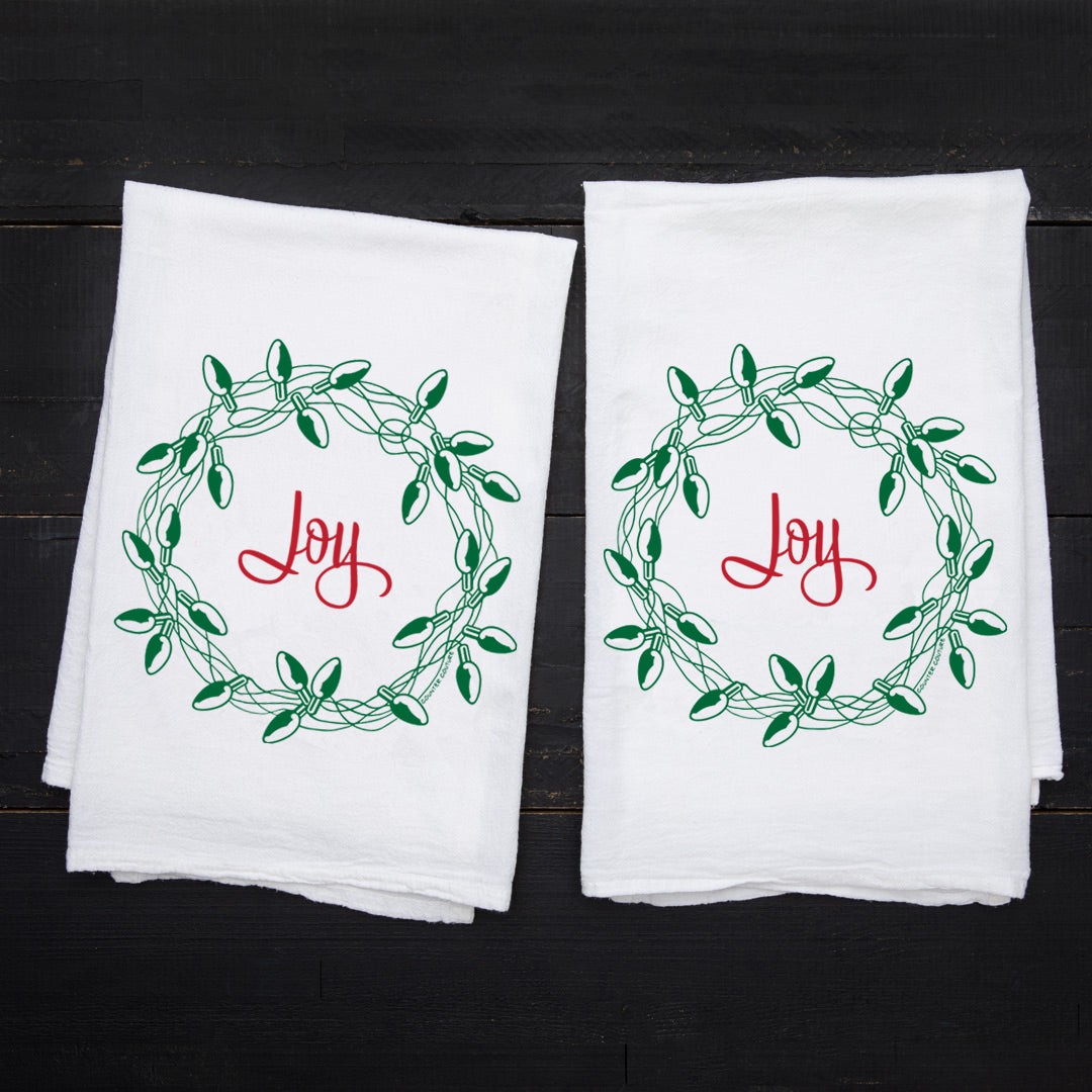 Set of two Joy Wreath Kitchen Towels by Counter Couture, each displaying a green light bulb wreath and the word 'Joy' in red on white cotton.