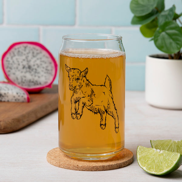 A 16oz Beer Can Glass featuring a hand-drawn illustration of a jumping baby goat, filled with a golden beverage, set on a cork coaster. Fresh lime slices are placed next to the glass, with a blurred background of colorful dragon fruit and a green plant.