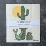 Counter Couture Swedish Dishcloth showcasing a design of a saguaro cactus and two smaller cacti with a yellow sun. The dishcloth is packaged with information about its absorbency and eco-friendly features.