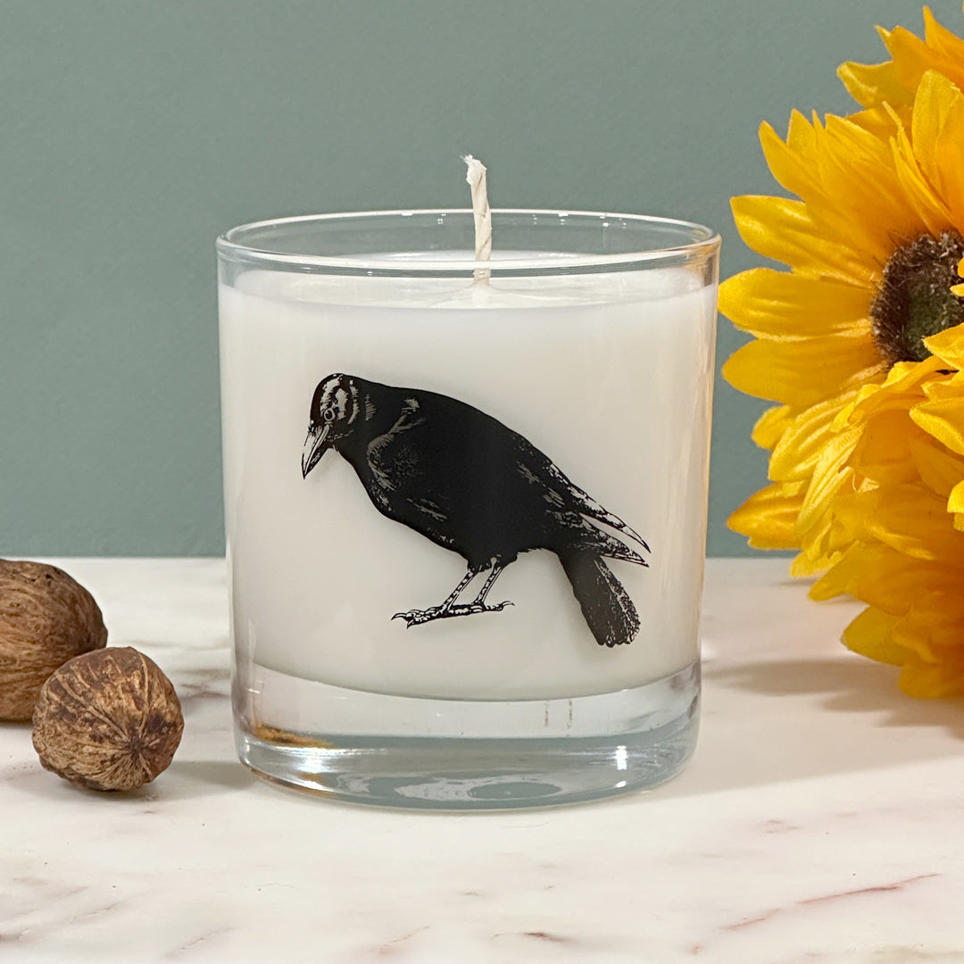 a candle with a black bird design on it, placed on a marble surface next to a yellow flower.