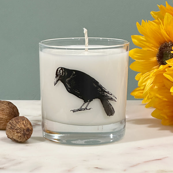 a candle with a black bird design on it, placed on a marble surface next to a yellow flower.