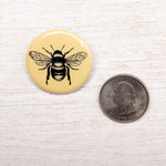 Yellow bee button pin next to a U.S. quarter on a light wood surface