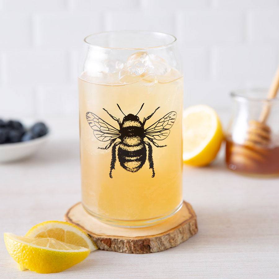 Bee can glass with iced drink, lemon slices, honey jar, and blueberries on a light table.