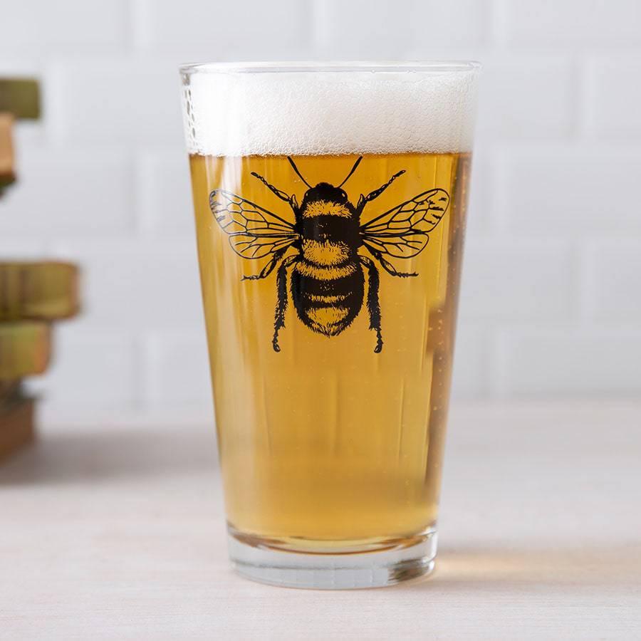 Pint glass of beer with black bee illustration on glass, white brick background