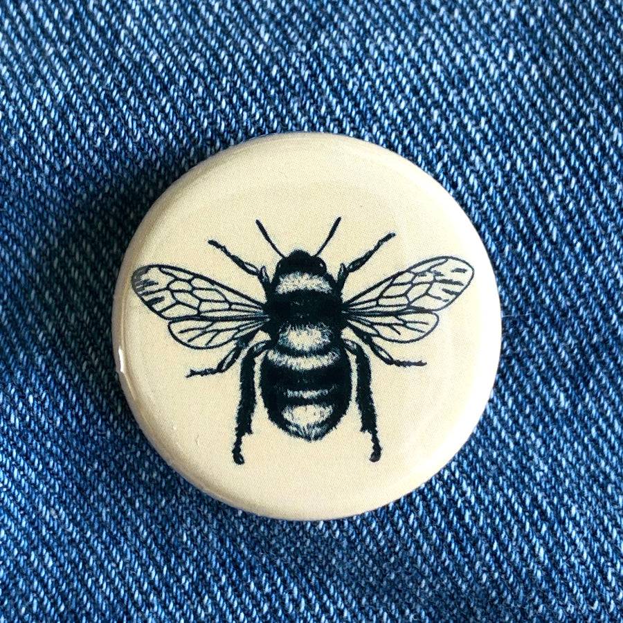 Round pin button with black bee illustration on cream background, placed on denim fabric