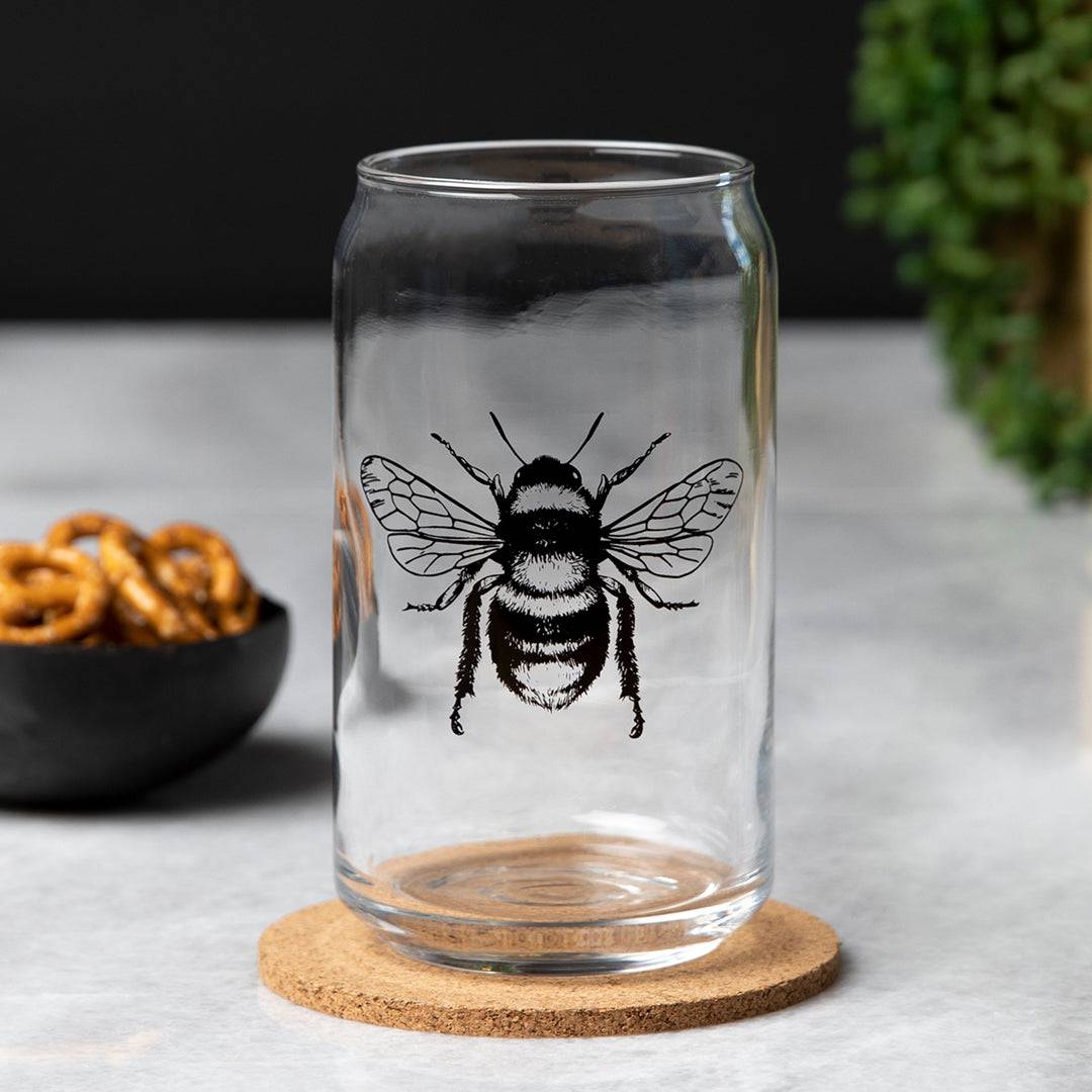 Clear can-shaped beer glass with black bee illustration on a cork coaster, modern barware