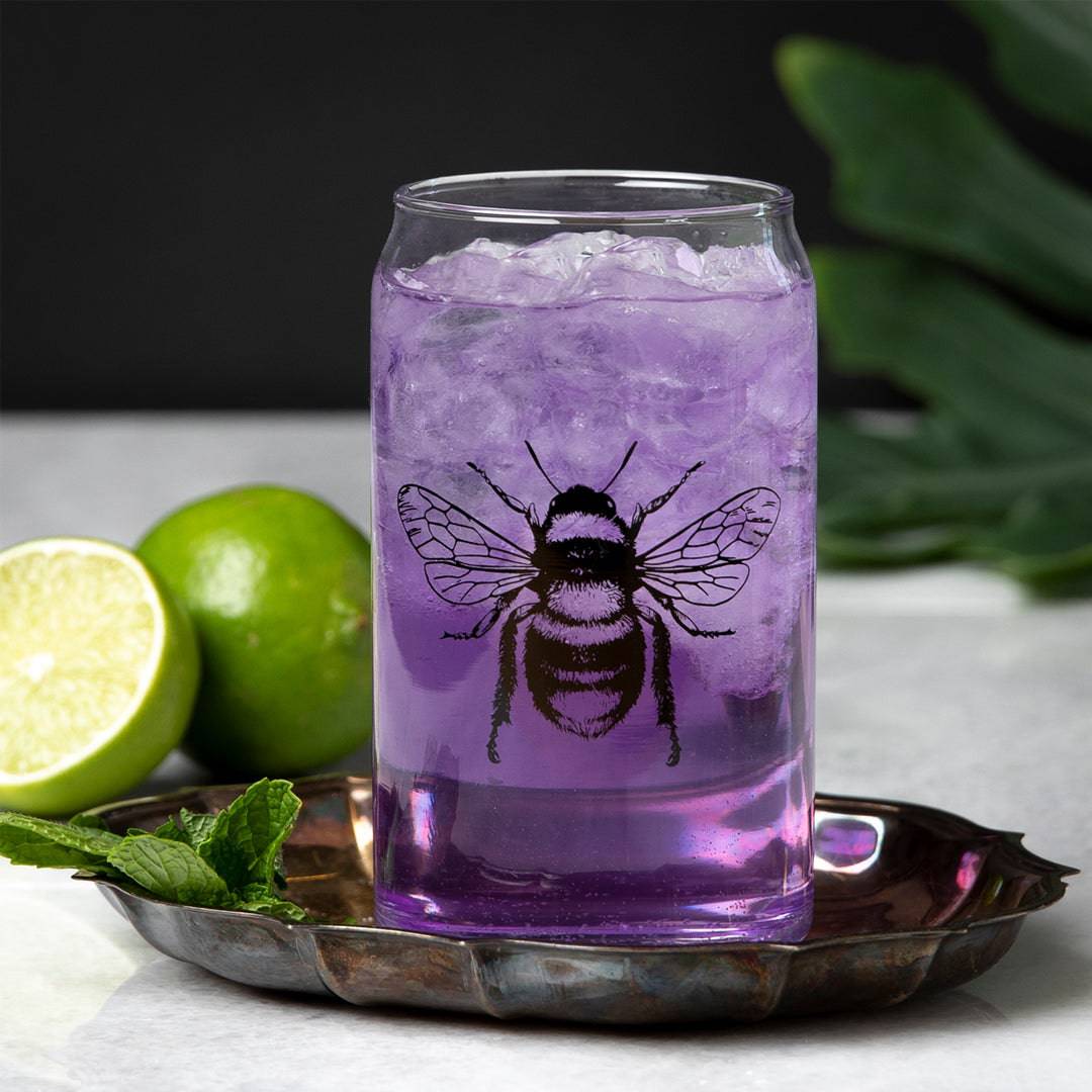 Bee can glass with purple drink and ice, garnished with lime and mint, on a tray.