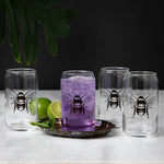 Set of four clear can-shaped bee glasses, one filled with purple drink, on marble surface