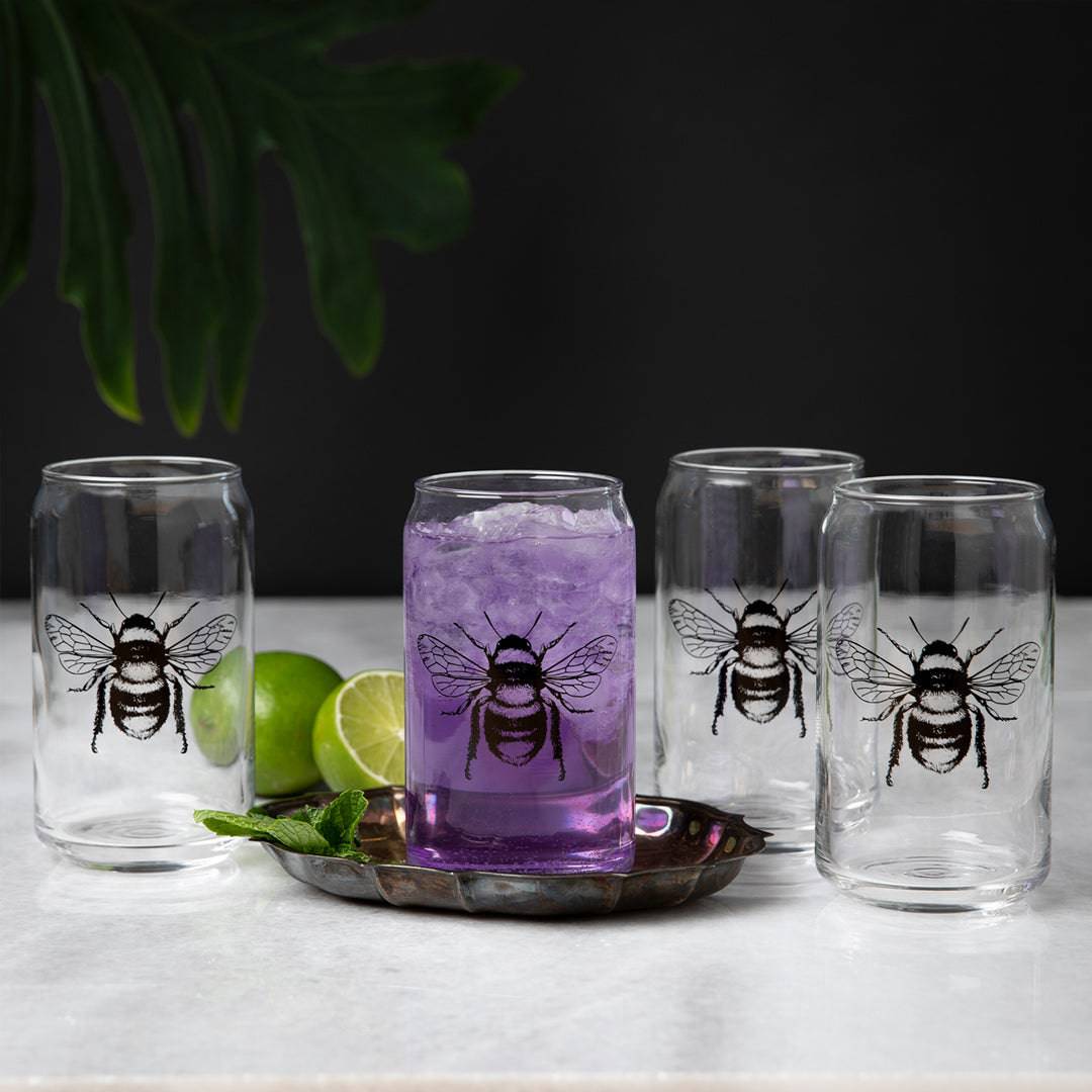 Set of four clear can-shaped bee glasses, one filled with purple drink, on marble surface