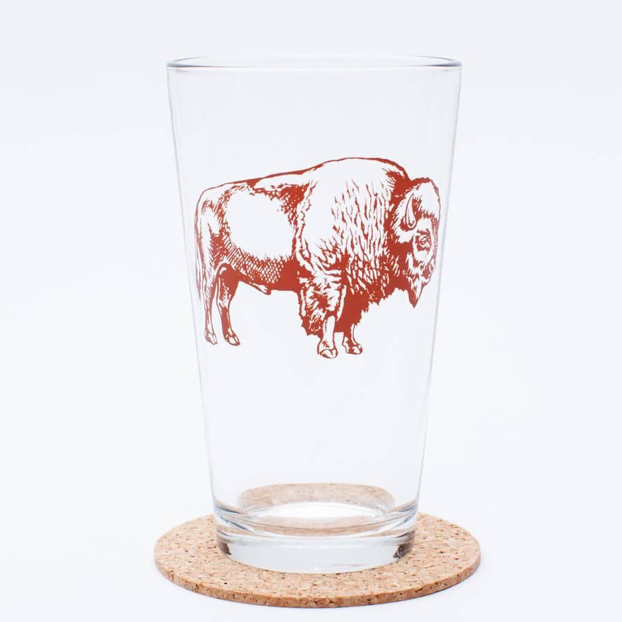 Clear pint glass with a detailed brown bison illustration, sitting on a cork coaster.