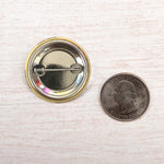 Metal pin-back button next to a US quarter on light wood surface for size comparison