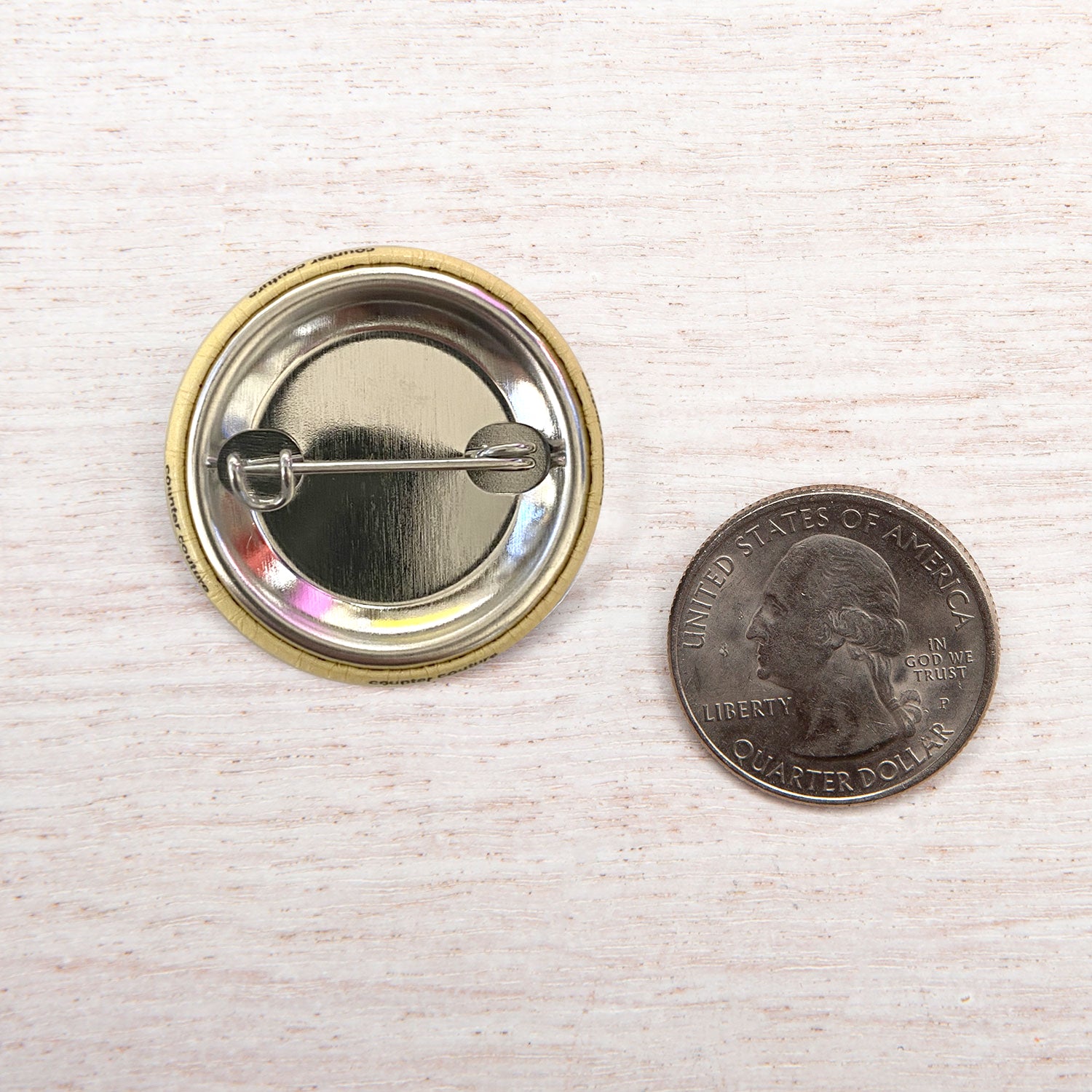 Metal pin-back button next to a US quarter on light wood surface for size comparison