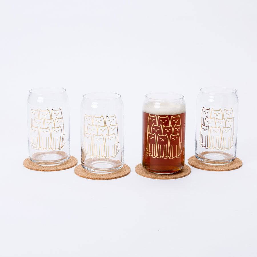 Set of four cat can glasses with gold cat illustrations, one filled with beer on cork coasters