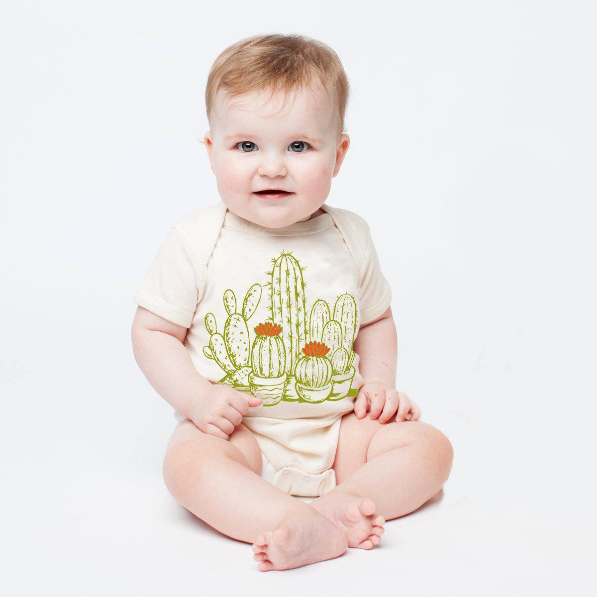 Baby wearing a cream onesie with green cactus and red flower graphic, sitting on white background.