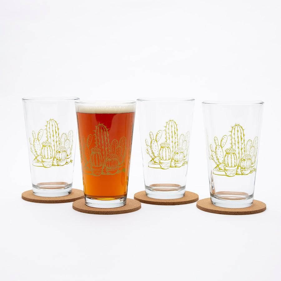 Set of four pint glasses with yellow cactus illustrations on cork coasters, one filled with beer.