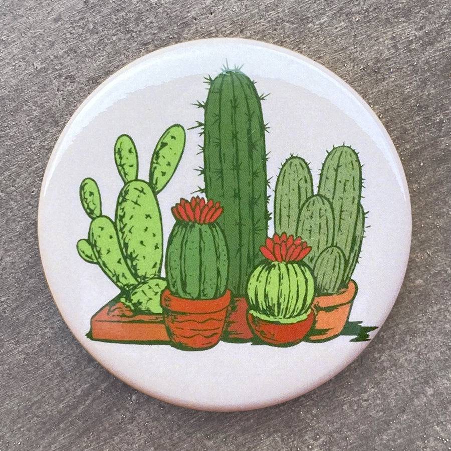 Round cactus magnet featuring illustrated green cacti in pots with red flowers on a gray background.