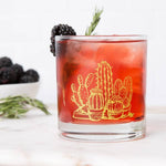 Whiskey glass with yellow cactus illustration holding a red cocktail, garnished with blackberry and rosemary