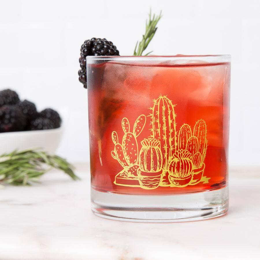 Whiskey glass with yellow cactus illustration holding a red cocktail, garnished with blackberry and rosemary