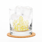 Whiskey glass with yellow cactus illustration, filled with ice, on a cork coaster