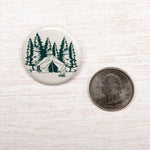 Round camping button with pine tree and tent design beside a US quarter on wood surface
