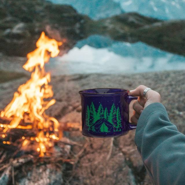Hand holding blue camping mug with forest and tent graphic near campfire by mountain lake