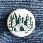 Camping button pin with tent, campfire, and pine trees illustration on denim fabric
