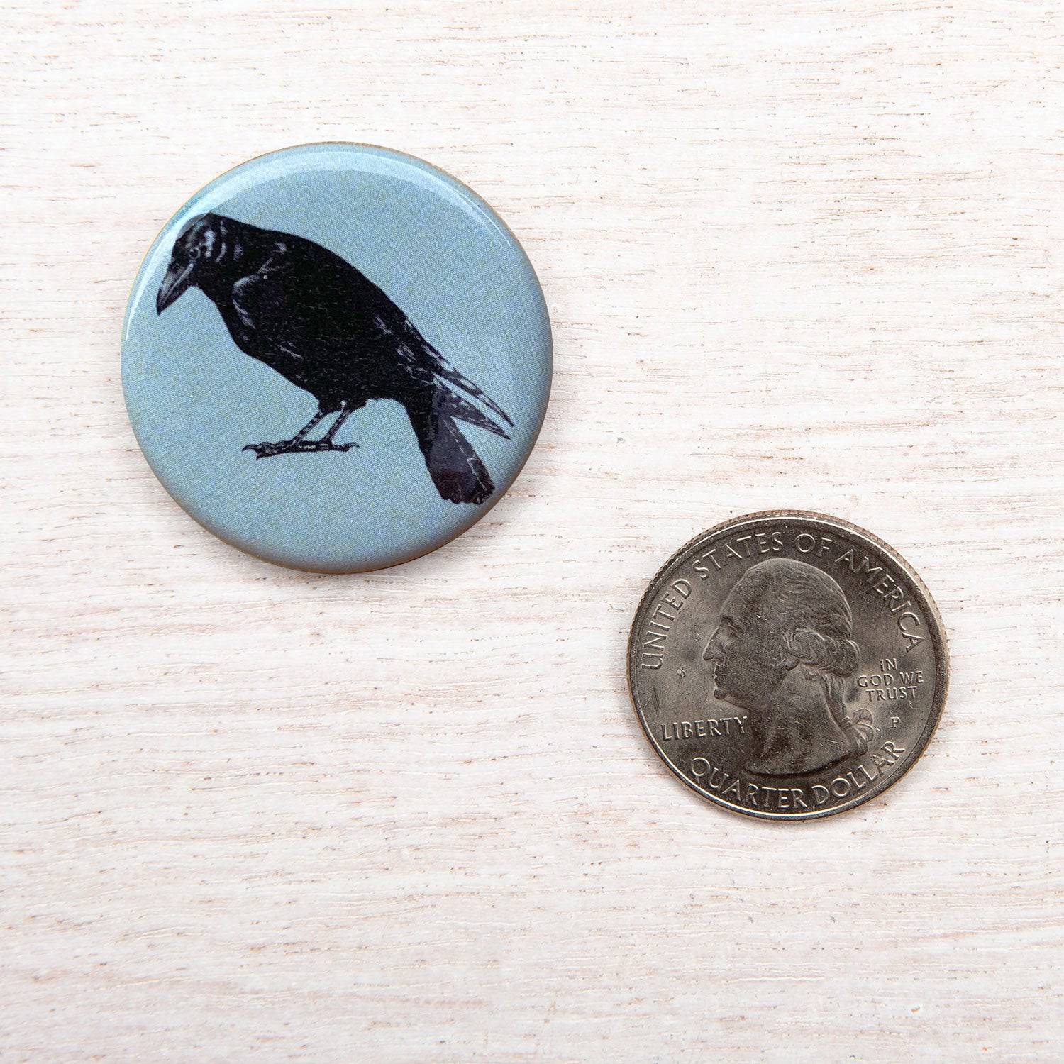 Round button with black crow illustration next to a US quarter on light wood surface