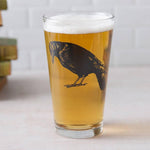 Pint glass of beer with black crow illustration, unique barware on light surface