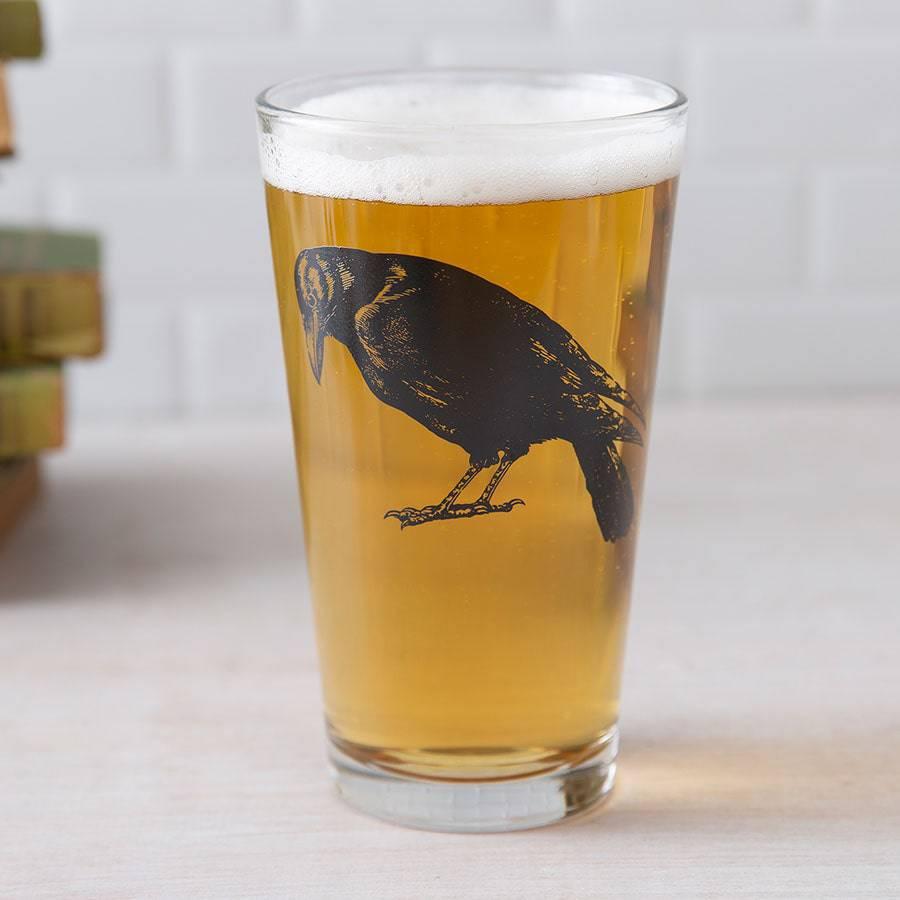 Pint glass of beer with black crow illustration, unique barware on light surface