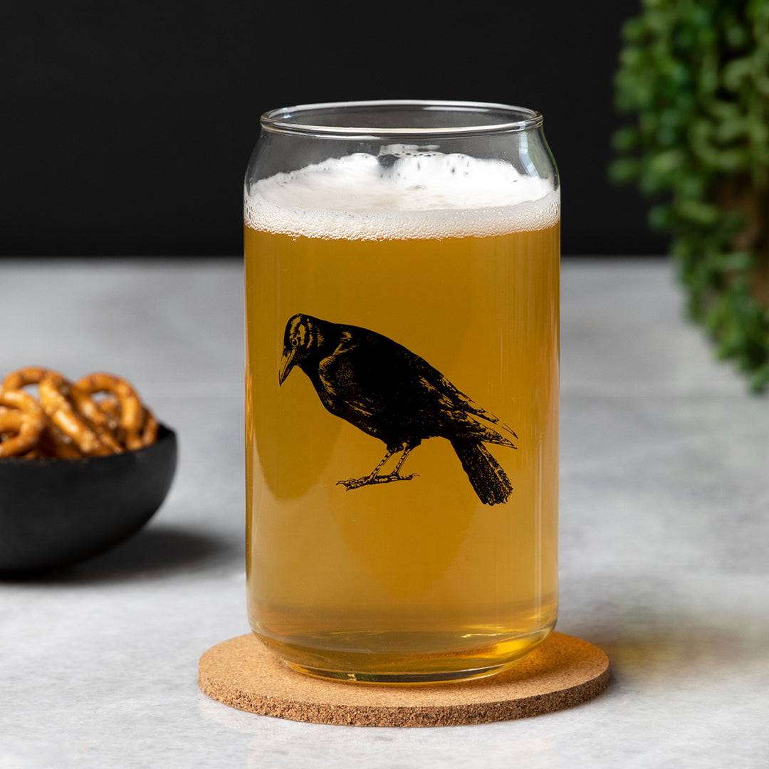 Can-shaped beer glass with black crow illustration, filled with beer on a cork coaster