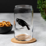 Clear can-shaped beer glass with black crow illustration, on a cork coaster