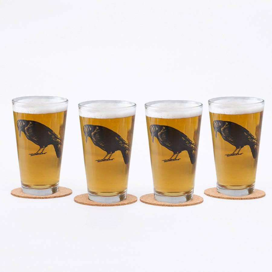 Set of four pint glasses with black crow illustration, filled with beer on coasters