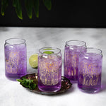 Set of four can-shaped glasses with gold dog illustrations, filled with purple drinks, on marble table.