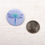 Blue dragonfly illustration button pin next to a US quarter on a light wood surface