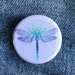 Round dragonfly button pin with blue and green insect illustration on denim fabric background