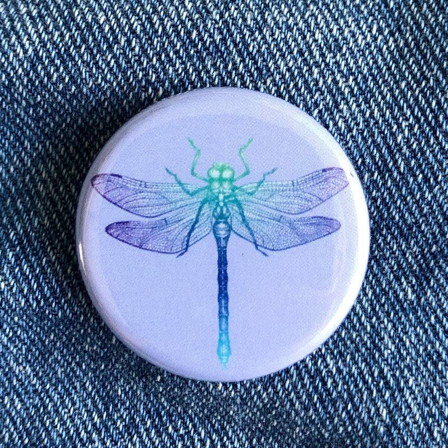 Round dragonfly button pin with blue and green insect illustration on denim fabric background
