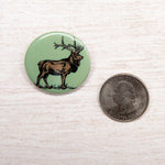 Green elk button pin with hand-drawn elk design, shown next to US quarter for size comparison