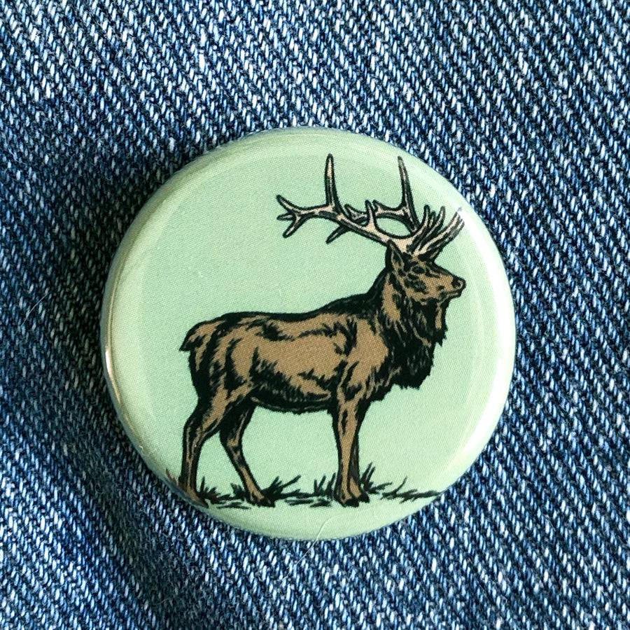 Round elk illustration button pin on denim fabric, rustic nature-themed accessory
