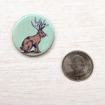 Round button with jackalope illustration beside a US quarter on light wood surface