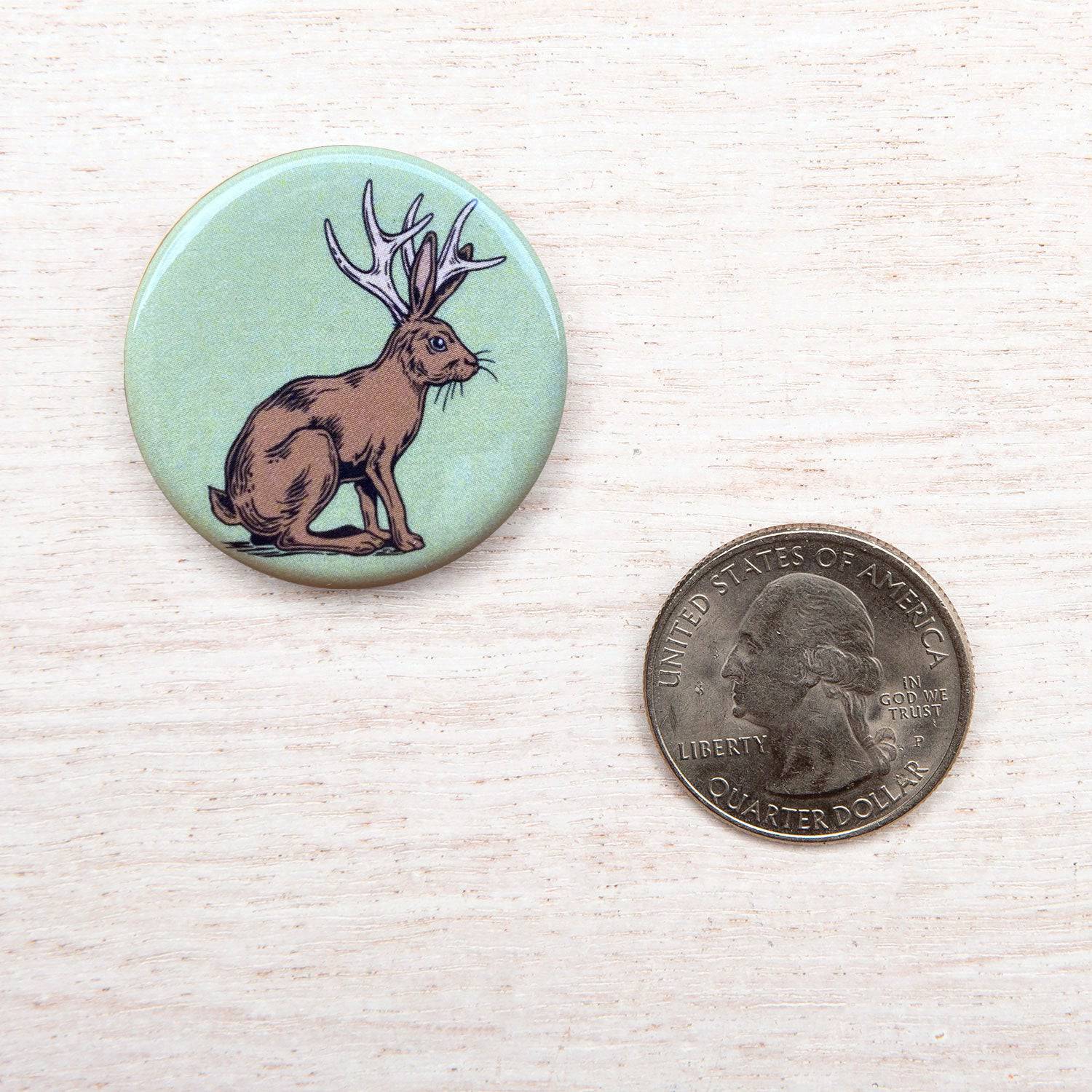 Round button with jackalope illustration beside a US quarter on light wood surface