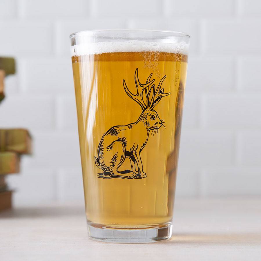 Pint glass with jackalope illustration, filled with beer, set on a light surface