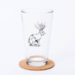 Pint glass with black jackalope illustration on a cork coaster, isolated on white background