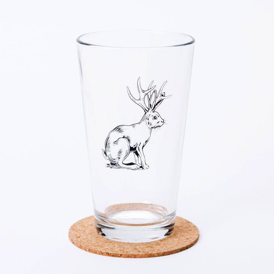 Pint glass with black jackalope illustration on a cork coaster, isolated on white background